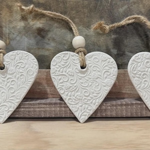 May include: Three white heart-shaped ornaments with a detailed floral pattern. Each heart is suspended by a natural twine cord and a wooden bead. The ornaments are displayed against a rustic wooden background.