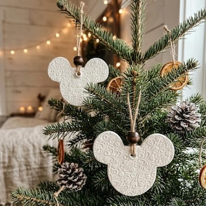 May include: White ceramic Mickey Mouse-shaped Christmas ornaments with a detailed floral pattern hang from a Christmas tree. The ornaments are suspended by twine and wooden beads. The tree is decorated with pinecones and dried orange slices.