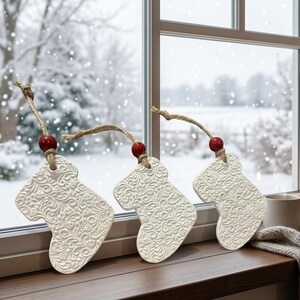 May include: Three white ceramic stocking ornaments featuring an intricate embossed floral pattern. Each ornament hangs from a natural jute twine loop accented with a single polished red wooden bead, creating a festive and textured appearance.