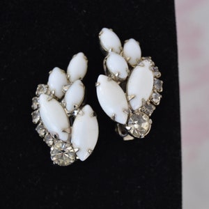 May include: A pair of white and clear rhinestone clip-on earrings. The earrings are shaped like a cluster of leaves with a teardrop-shaped center stone.