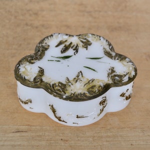 May include: A white porcelain trinket box with a decorative, ornate, gold-painted design. The box has a clover-shaped design with green painted leaves.