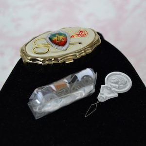 May include: A gold metal sewing kit with a white enamel lid featuring a colorful shield with a sewing needle and scissors. The kit includes a small package of needles, thread, and spools.