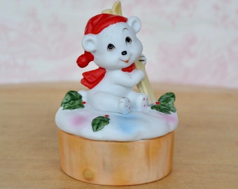 May include: A ceramic figurine of a white polar bear wearing a red hat and scarf, sitting on a snow-covered base decorated with green holly leaves and red berries. The bear holds a pair of yellow skis, and the base is a tan cylindrical shape.