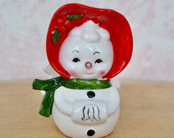 May include: A ceramic snowman figurine featuring a glossy white finish, a bright red bonnet adorned with green holly leaves and red berries, a dark green scarf, and two black buttons on the torso. The snowman has painted facial features.