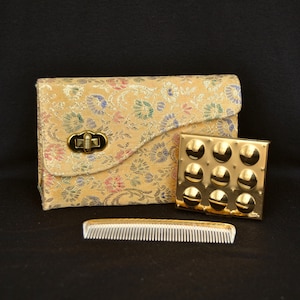 May include: A vintage gold-colored compact mirror with a floral patterned case. The case is made of a brocade fabric with a gold clasp. A comb is also included.