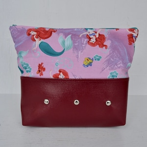 May include: A red zippered pouch with a pink fabric lining featuring a repeating pattern of Ariel the Little Mermaid and Flounder. The pouch has three silver rivets on the front.