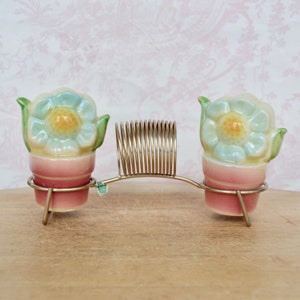 May include: A set of two ceramic salt and pepper shakers shaped like flower pots with blue flowers. The shakers are on a gold metal stand with a spring.