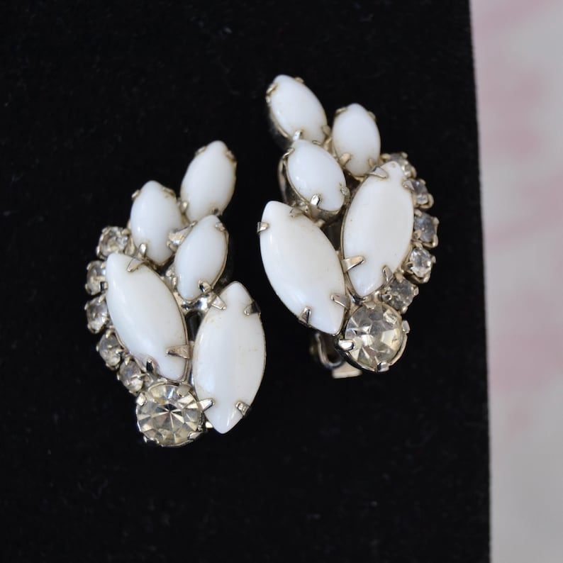 May include: A pair of white and clear rhinestone clip-on earrings. The earrings are shaped like a cluster of leaves with a teardrop-shaped center stone.