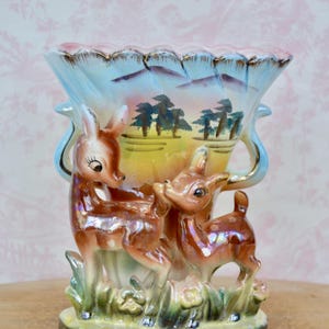 May include: A vintage ceramic vase with a light blue and pink rim, featuring two deer figures and a painted landscape scene. The vase has a flared shape with handles and is decorated with gold accents.