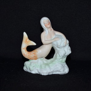 May include: A porcelain figurine of a mermaid with white hair and a peach-colored tail. The mermaid is seated on a light green base, holding a white cloud-like form. The tail has orange and peach details.