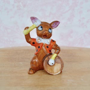 Puede incluir: Figura de cerámica marrón de un conejo con chaqueta roja de lunares negros y pajarita blanca, tocando un tambor. El conejo sostiene baquetas amarillas, una levantada. Ojos azules, orejas rosas, una encantadora pieza decorativa.