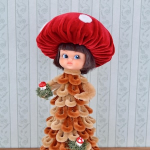 May include: A vintage mushroom doll with a red velvet cap featuring white spots. The doll's dress is made of looped brown and tan fabric. It holds a small mushroom and is a whimsical decorative item.