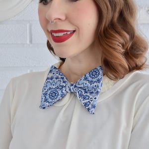 May include: A blue and white patterned bow tie. The bow tie features a repeating floral design in shades of blue and white. The bow tie is worn with a white collared shirt.