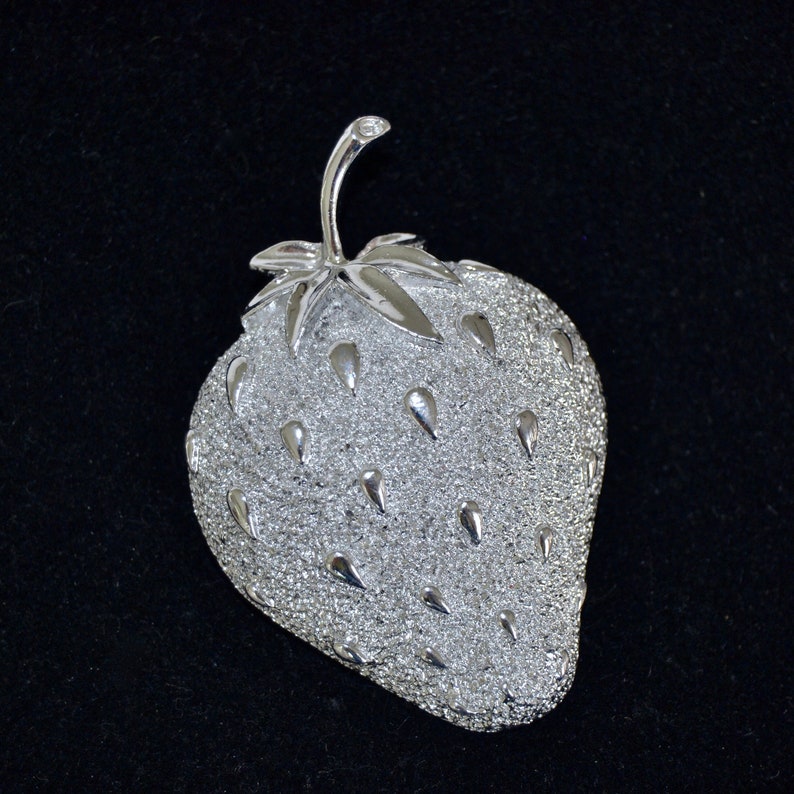Pu&ograve; includere: Un broche a forma di fragola argentato con una superficie testurizzata. Il broche ha uno stelo e foglie.