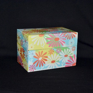 May include: A rectangular box with a hinged lid, decorated with a repeating pattern of large daisies in shades of pink, orange, white, and yellow against a light blue background. The box is likely made of cardboard or a similar material.