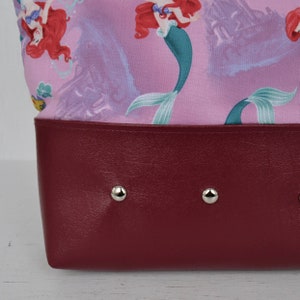 Puede incluir: Una bolsa de tela rosa con un estampado de Ariel, la Sirenita, con un fondo de cuero burdeos y remaches plateados.