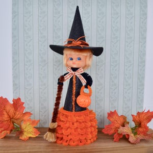May include: A Halloween witch doll with a black hat, dress, and broom. The doll has a blonde wig, blue eyes, and an orange pumpkin-shaped treat bag. The skirt is made of orange chenille. Autumn leaves are placed around the doll.