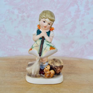 May include: A vintage porcelain figurine of a young girl holding a broom, wearing a green and white dress. A small squirrel and a basket are at her feet. The figurine has a cream-colored base and is set against a pink floral background.