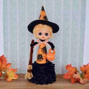 May include: A Halloween-themed doll dressed as a witch. The doll has blonde hair, blue eyes, and wears a black hat and dress. The orange cone-shaped body is decorated with black bats and a bow. The doll holds a broom and a carved pumpkin.