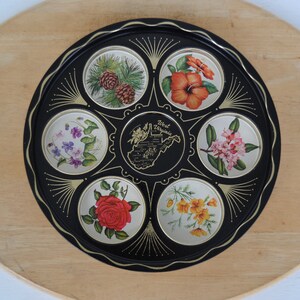 May include: A black serving tray with a gold rim and decorative accents. The tray features circular images of flowers and pinecones, with a central image of the West Virginia state map and text.