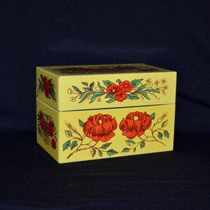 May include: A yellow rectangular box with a lid, decorated with red roses and green leaves. The box has a floral design on the top and front, set against a dark blue background.