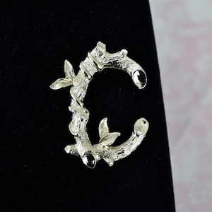 May include: A gold-toned metal brooch shaped like the letter C, formed from a stylized branch with leaves.