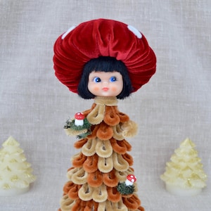 May include: A doll with a dark red velvet mushroom cap hat featuring white spots, black hair, and blue eyes. The body is constructed from layered tan and brown pipe cleaners, decorated with two small red and white mushroom ornaments.