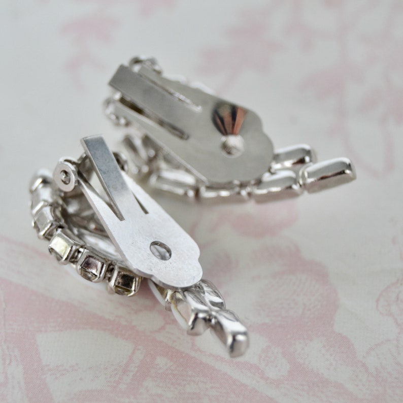 May include: A pair of silver tone clip-on earrings with clear rhinestones. The earrings are in the shape of a flower with a cluster of rhinestones in the center.