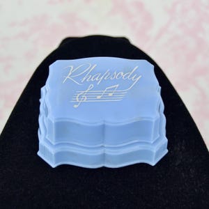 May include: A light blue, scalloped-edge box with the word "Rhapsody" and musical notes in white lettering on the lid. The box is sitting on a black surface, with a pink and white background.