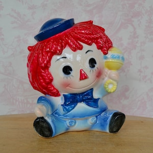 May include: A ceramic piggy bank shaped like a rag doll with red hair, a blue sailor hat, and a blue and white outfit. The doll is holding a yellow rattle with blue and white accents.