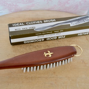 May include: A brown wooden clothes brush with a gold fleur-de-lis design and a gold ring handle. The brush has white bristles and is packaged in a box that reads "Ideal Clothes Brush with Shoe Horn".