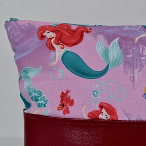 Puede incluir: Una bolsa de tela rosa con cremallera y fondo de cuero rojo. La tela presenta un patr&oacute;n repetido de Ariel, la Sirenita, con su cola y un castillo en el fondo.