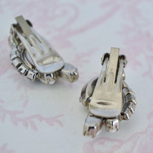 May include: A pair of silver tone clip-on earrings with clear rhinestones. The earrings are in the shape of a flower with a round center.