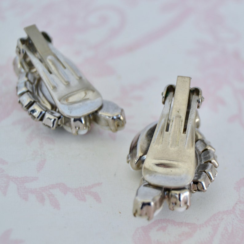 May include: A pair of silver tone clip-on earrings with clear rhinestones. The earrings are in the shape of a flower with a round center.