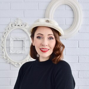 Peut inclure: Un chapeau vintage crème avec un nœud et un bijou décoratif. Le chapeau est porté par une personne avec du rouge à lèvres rouge et des cheveux bruns. L'arrière-plan présente des cadres décoratifs blancs et un mur de briques blanches.