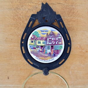 May include: A black metal horseshoe-shaped wall hanging with a ceramic center featuring a colorful illustration of a building and people. The text "Old Coach House Bristol" is visible. A gold-colored ring is attached at the bottom.