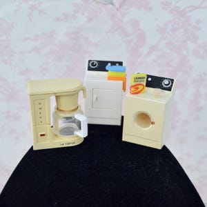May include: A miniature beige coffee maker with a glass carafe, a white dryer with stacked colorful towels, and a beige washing machine with a small box of laundry detergent on top. The coffee maker features a numbered scale and the text Le Coffee.