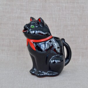 May include: A glossy black ceramic creamer shaped like a sitting cat. The cat features a bright red painted scarf around its neck, vibrant green eyes, and red accents inside the ears. A curved handle is attached to the back of the cat figure.
