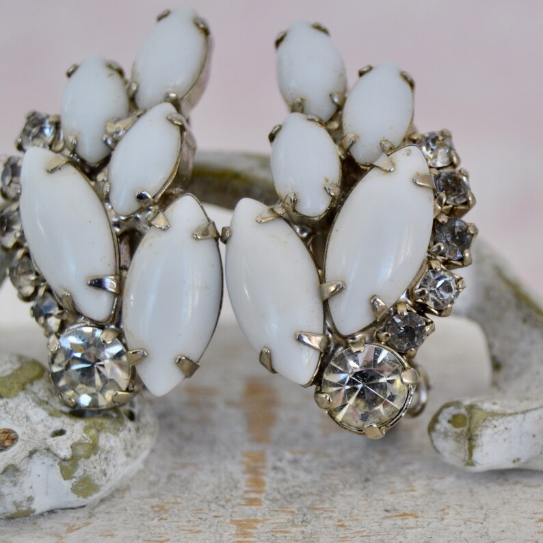 May include: A pair of vintage clip-on earrings featuring a cluster of white oval-shaped stones surrounded by clear rhinestones.