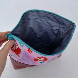 Puede incluir: Una bolsa de cremallera rosa con fondo rojo y cremallera turquesa. La bolsa est&aacute; forrada con un tejido azul con lunares blancos. La parte delantera de la bolsa presenta una impresi&oacute;n de Ariel de La Sirenita.