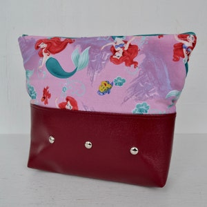 Puede incluir: Una bolsa de cremallera rosa y roja con un estampado de Ariel la Sirenita y Sebasti&aacute;n. La bolsa tiene un fondo burdeos con tachuelas plateadas.