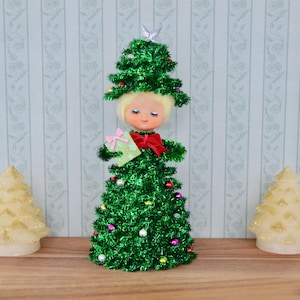 May include: A charming Christmas tree ornament featuring a doll's face, crafted from green tinsel and decorated with colourful baubles. It holds a small gift with a pink bow and wears a red bow tie. A silver star sits atop the tree, perfect for festive decor.