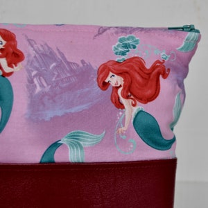Puede incluir: Bolsa de cremallera de tela rosa con fondo de cuero rojo. La bolsa presenta un patr&oacute;n repetido de Ariel, la Sirenita, con un castillo en el fondo.