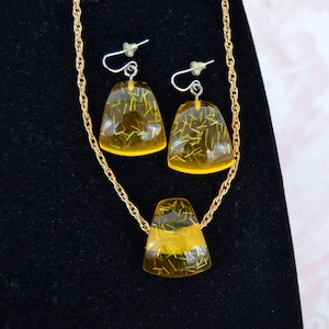 May include: A gold chain necklace and matching earrings with yellow resin pendants. The pendants have a glittery, green and gold, speckled appearance.