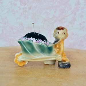 May include: A ceramic figurine of a child pushing a green and yellow wheelbarrow that serves as a pincushion. The wheelbarrow features a dark green velvet cushion adorned with small pink floral accents and two silver sewing pins inserted.