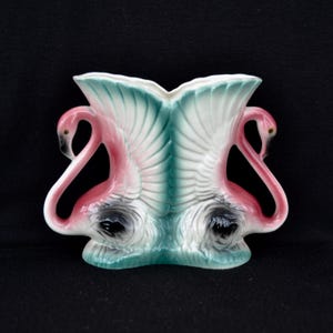 May include: A ceramic vase featuring a flamingo design. The vase is shaped with two flamingo figures facing each other, with pink bodies, black beaks, and teal and white wings. The vase has a glossy finish and is set against a black background.