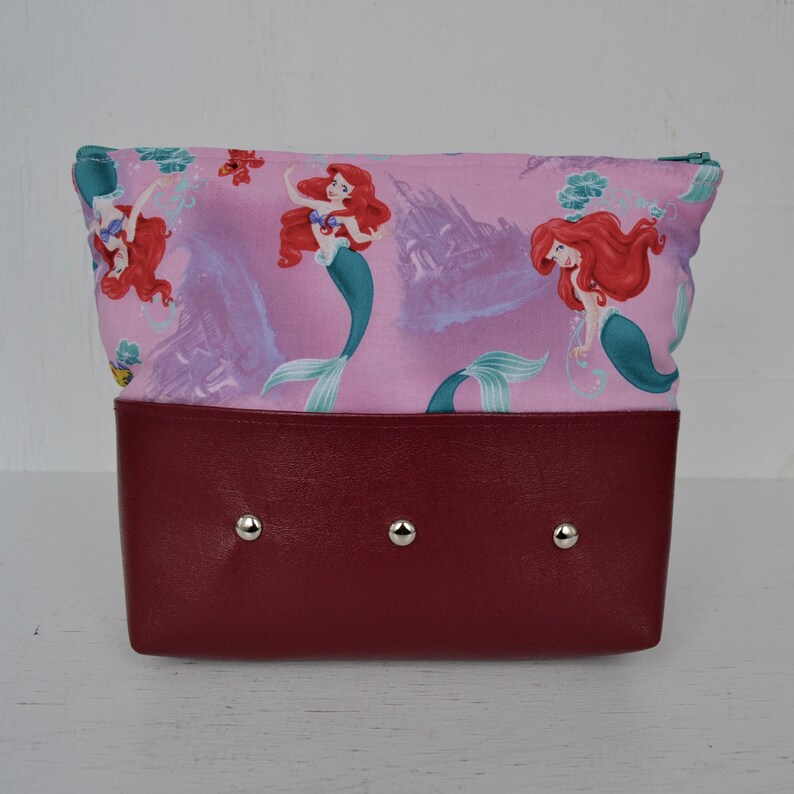 Puede incluir: Una bolsa rosa con cremallera y fondo burdeos. La bolsa presenta un patr&oacute;n repetido de Ariel la Sirenita en varias poses. La bolsa tiene tres remaches plateados en la parte inferior.