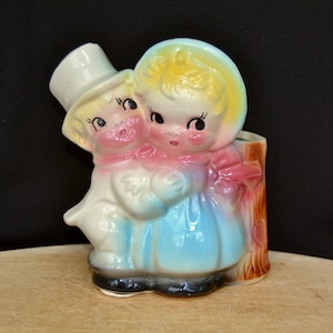 May include: A vintage ceramic planter shaped like a boy and girl holding hands. The boy is wearing a top hat and the girl is wearing a blue dress and a pink bow. The planter has a brown wooden log behind them.