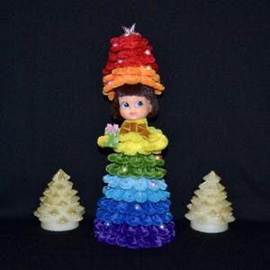 May include: A doll in a rainbow Christmas tree costume, holding a gift. The dress is a gradient of colours, from red to purple, with pearl accents. Two gold glittery Christmas tree candles flank the doll.