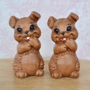 May include: A pair of brown ceramic bunny salt and pepper shakers. The bunnies have big eyes, a pink tongue sticking out, and are holding their paws in front of their faces.